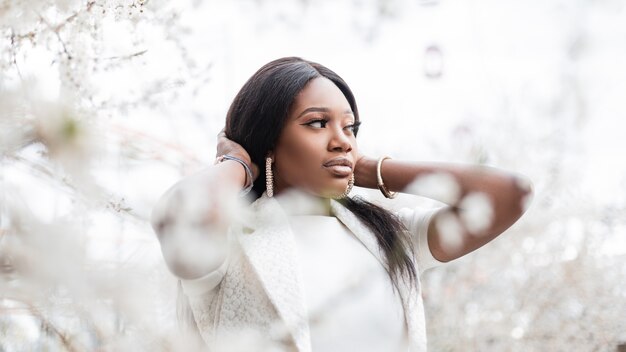 Mujer africana negra de moda bastante joven en un parque florecido camina en la naturaleza. Chica negra en flores blancas de primavera al aire libre