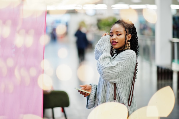 Mujer africana posando en suéter y jeans posando en el centro comercial. Bokeh de Navidad y año nuevo tema.