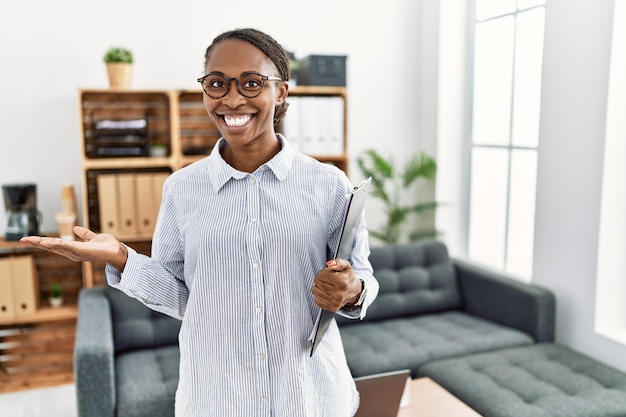 Mujer africana que trabaja en la clínica de psicología señalando a un lado con las manos abiertas mostrando espacio de copia, presentando un anuncio sonriendo emocionada feliz