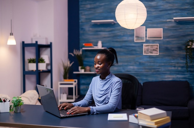 Mujer africana que trabaja de forma remota desde la oficina en casa por la noche en la computadora portátil. Empresario negro sentado en el lugar de trabajo personal escribiendo en el teclado.