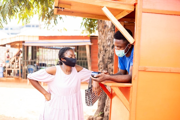 Mujer africana en un quiosco de servicio pos recogiendo algo de dinero para un cliente, usa una máscara facial