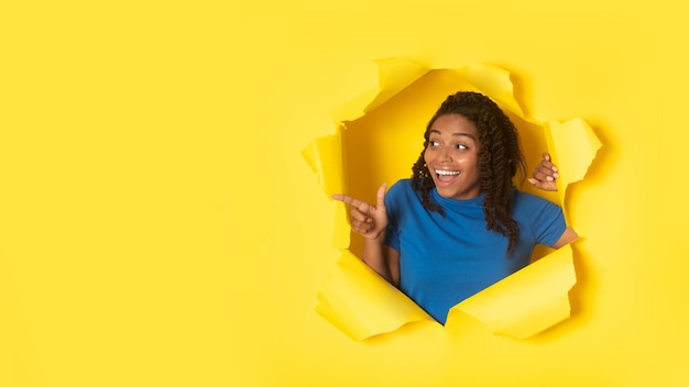 Mujer africana señalando con el dedo a un lado a través del fondo de papel amarillo roto