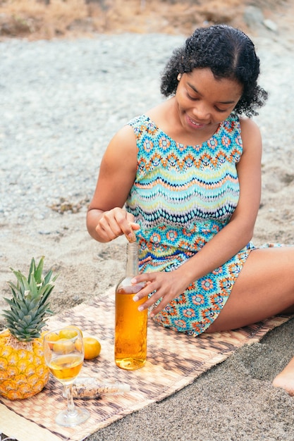 Mujer africana sentada en la playa descorchando una botella de vino