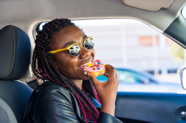 Mujer africana sonriendo mientras muerde una rosquilla dentro de un coche