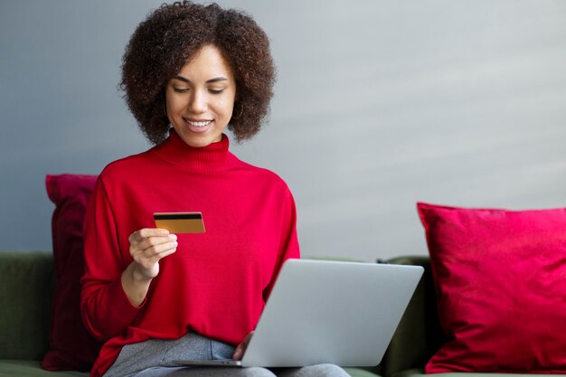 Mujer africana sonriente sosteniendo una tarjeta de crédito usando una computadora portátil para comprar en línea buscando el saldo de la tarjeta