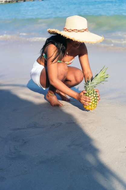 Mujer africana sostiene una piña con el mar de fondo.