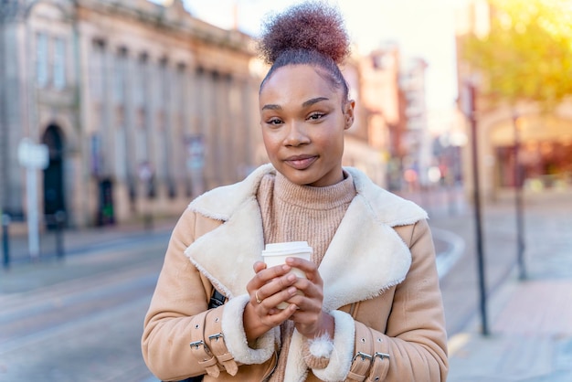 Mujer africana sostiene una taza de café Una mujer morena rizada sonriente con suéter marrón esperando un tranvía