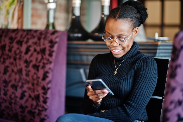 Mujer africana en suéter negro sentado en el café y mirar el teléfono móvil.