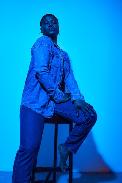 Mujer africana de talla grande y segura con camisa de mezclilla y pantalones posando en una silla en el estudio con fondo de neón azul