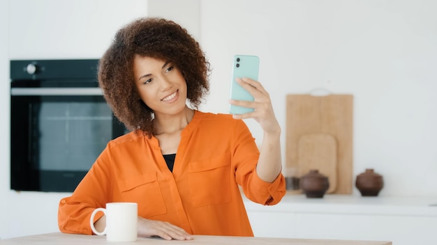 Mujer africana con vlogger femenina de cabello rizado en la cocina en casa tomando fotos en el teléfono móvil