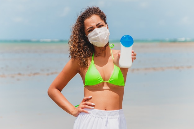 Mujer afro en bikini y máscara con botella de protector solar en la mano en la playa.