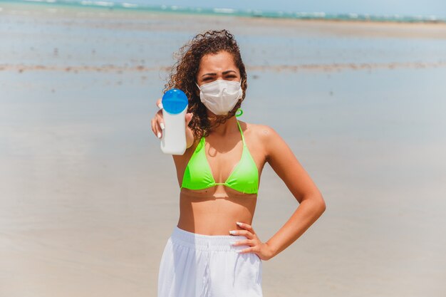 Mujer afro en bikini y máscara con botella de protector solar en la mano en la playa.