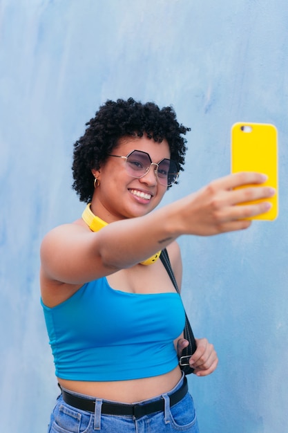 Mujer afro caminando por la calle y tomando un selfie con su teléfono celular, sonriendo.