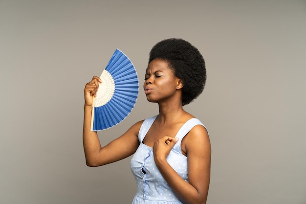 Mujer afro disgustada sufre de calor dentro de la mujer africana sobrecalentada saludando con ventilador de papel