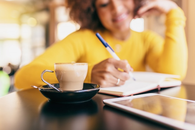 Mujer afro joven en el café. Ella está escribiendo en un cuaderno, primer plano de un café.