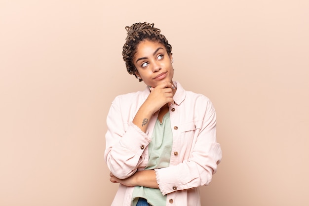 Mujer afro joven que se siente pensativa, preguntándose o imaginando ideas, soñando despierto y mirando hacia arriba para copiar el espacio