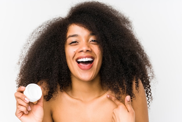 La mujer afro joven que sostenía una crema hidratante aislada sorprendió señalar a sí mismo, sonriendo ampliamente.