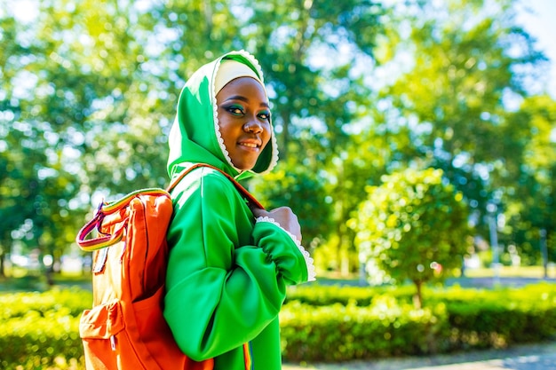 Mujer afro moderna en hiyab verde con maquillaje brillante y mochila holg orabge de nariz penetrante en el parque