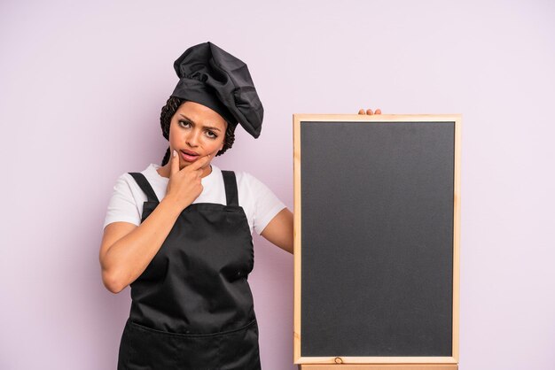 Mujer afro negra con la boca y los ojos bien abiertos y la mano en la barbilla chef y concepto de pizarra