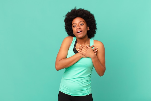 Mujer afro negra que se siente conmocionada y sorprendida, sonriendo, tomando la mano al corazón, feliz de ser la indicada o mostrando gratitud