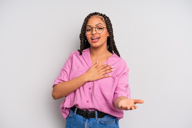 Mujer afro negra que se siente feliz y enamorada, sonriendo con una mano al lado del corazón y la otra estirada al frente