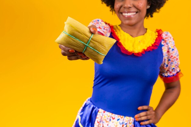 Mujer afro en ropa de festa junina sosteniendo un tamal sobre fondo amarillo