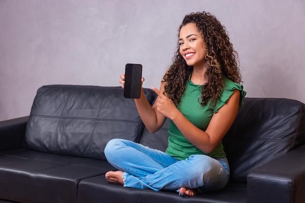 Mujer afro sentada en el sofá sosteniendo el teléfono celular con pantalla en blanco con espacio libre para texto.