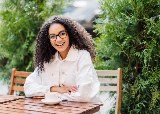 Mujer afro vestida de blanco, bebe café en la cafetería al aire libre