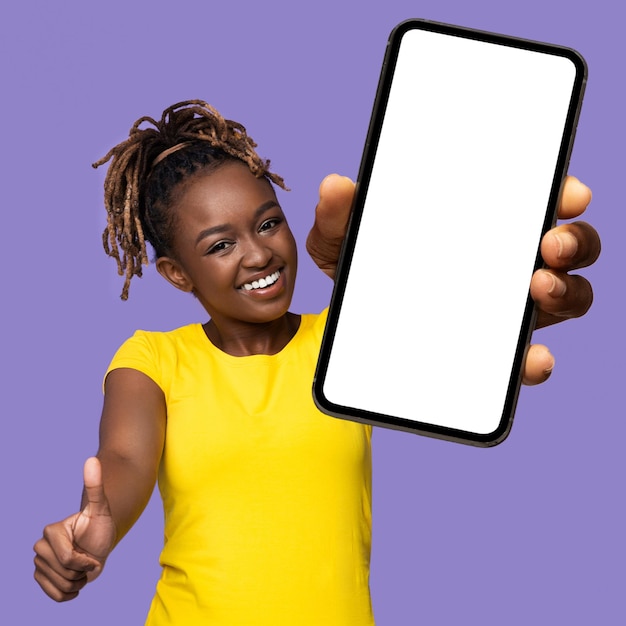 Mujer afroamericana alegre que muestra el teléfono celular y el pulgar hacia arriba