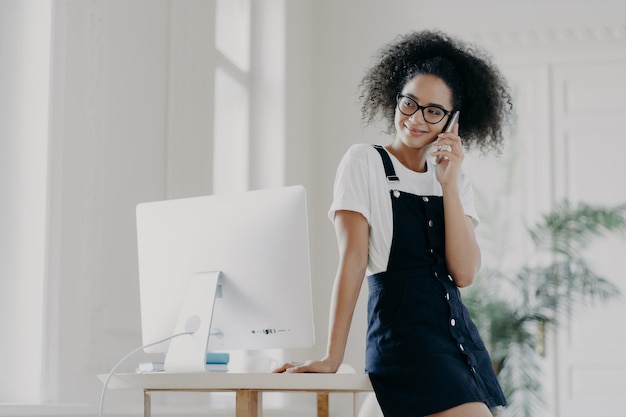 Una mujer afroamericana de aspecto agradable tiene una conversación telefónica que pide consejo antes de una entrevista de trabajo vestida con ropa elegante posa cerca del lugar de trabajo con tecnología moderna Comunicación