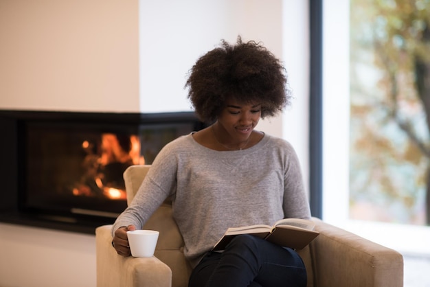 mujer afroamericana bebiendo una taza de café leyendo un libro en la chimenea. Joven negra con bebida caliente relajante calentamiento calentamiento. otoño en casa.