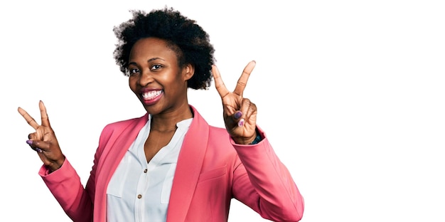 Mujer afroamericana con cabello afro con chaqueta de negocios sonriendo con la lengua afuera mostrando los dedos de ambas manos haciendo el signo de la victoria número dos