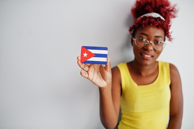 Mujer afroamericana con cabello afro usa camiseta amarilla y anteojos sostiene la bandera de Cuba aislada sobre fondo blanco