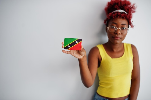 Mujer afroamericana con cabello afro usa camiseta amarilla y anteojos sostiene la bandera de San Cristóbal y Nieves aislada en fondo blanco