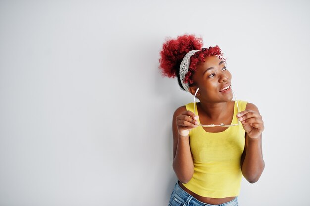 Mujer afroamericana en camiseta amarilla y anteojos contra la pared blanca.