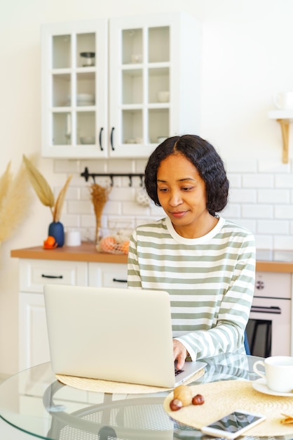 Foto mujer afroamericana centrada en el trabajo remoto mientras está sentada en la cocina y usando una computadora portátil