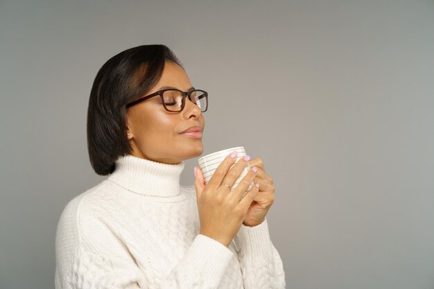 Mujer afroamericana complacida en suéter caliente disfrutando de una deliciosa bebida caliente de invierno con los ojos cerrados