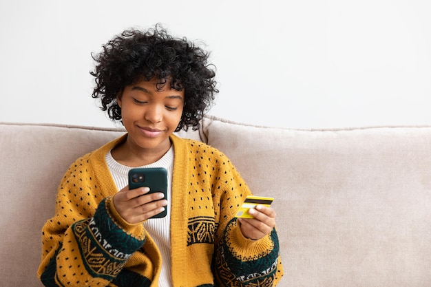 Mujer afroamericana comprando en línea con un teléfono inteligente pagando con tarjeta de crédito dorada Chica sentada en casa comprando en Internet ingrese los detalles de la tarjeta de crédito Servicio de entrega de comercio electrónico de compras en línea