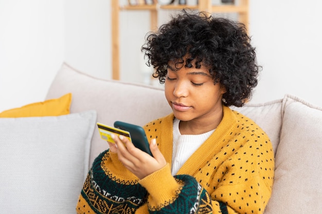 Mujer afroamericana comprando en línea con un teléfono inteligente pagando con tarjeta de crédito dorada Chica sentada en casa comprando en Internet ingrese los detalles de la tarjeta de crédito Servicio de entrega de comercio electrónico de compras en línea