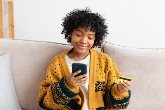 Mujer afroamericana comprando en línea con un teléfono inteligente pagando con tarjeta de crédito dorada Chica sentada en casa comprando en Internet ingrese los detalles de la tarjeta de crédito Servicio de entrega de comercio electrónico de compras en línea