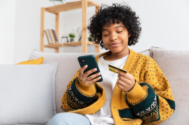 Mujer afroamericana comprando en línea con teléfono inteligente pagando con tarjeta de crédito de oro chica sentada