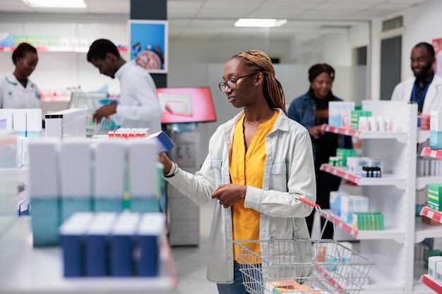 Mujer afroamericana comprando medicamentos en la farmacia, sosteniendo un paquete de tabletas. Cliente eligiendo píldoras, cliente de pie en el pasillo de la farmacia, negocio farmacéutico, concepto de venta minorista de medicamentos