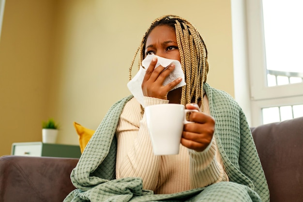 Mujer afroamericana cubierta con una manta que sopla la nariz que moquea sosteniendo una taza de té