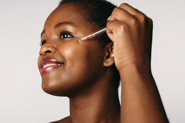 Mujer afroamericana dejando caer crema hidratante de colágeno en suero en su rostro aislado sobre fondo blanco...