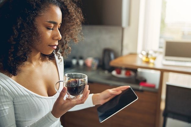 Mujer afroamericana delgada de pie en la cocina con su teléfono móvil mientras sostiene un vaso de café