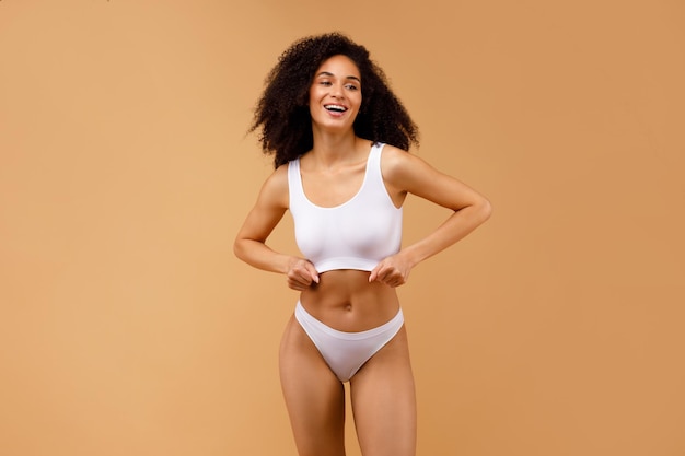 Mujer afroamericana delgada en sujetador superior de ropa interior blanca y bragas de pie aislado sobre beige