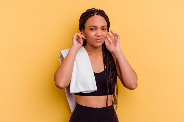 Mujer afroamericana de deporte joven sosteniendo una toalla aislada sobre fondo amarillo cubriendo las orejas con las manos.