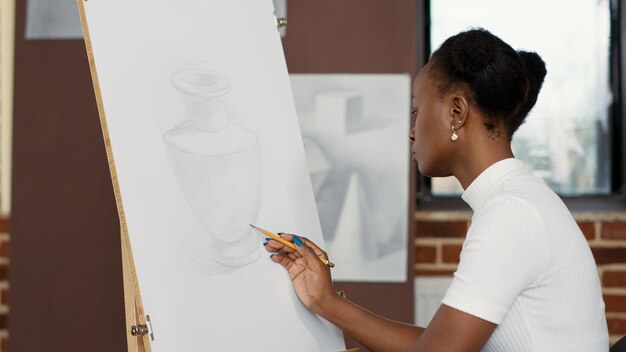 Mujer afroamericana dibujando modelo de jarrón sobre lienzo blanco para desarrollar habilidades creativas para el crecimiento educativo. Pasatiempo contemporáneo para practicar bocetos de clase de arte, aprender a dibujar objetos.