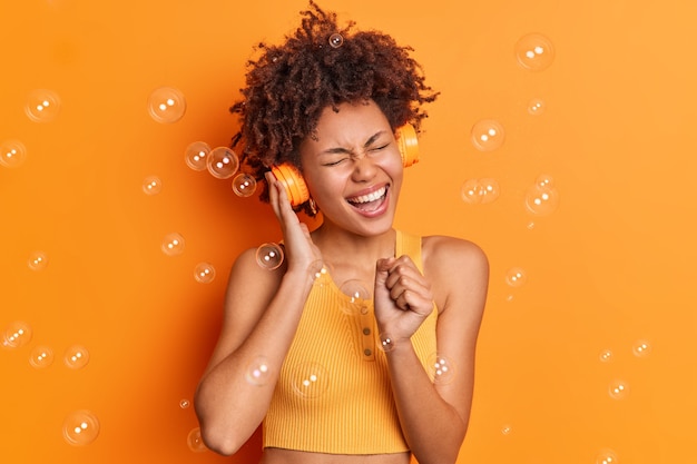 La mujer afroamericana divertida optimista canta la canción mantiene la mano cerca de la boca como si el micrófono usara auriculares inalámbricos escucha su música favorita aislada sobre una pared naranja con pompas de jabón