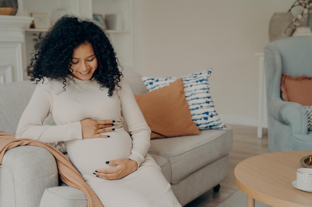 Mujer afroamericana embarazada tomando un descanso en el sofá en casa hablando con el futuro bebé